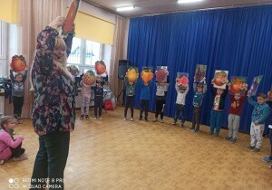 Dzieci stoją na dużej sali w kolorowych kapeluszach. Stoją jeden obok drugiego. Przed nimi stoi pani prowadząca koncert.