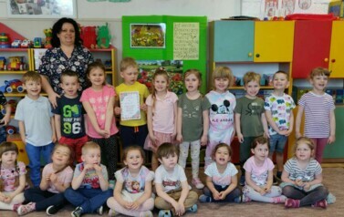 Zdjęcie grupowe dzieci w sali przedszkolnej. Uśmiechają się do zdjęcia. Na śrdoku chłopiec trzyma w rękach dyplom. Razem z nimi stoi nauczycielka.