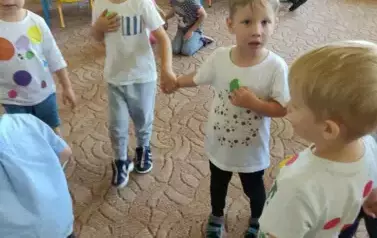 Dzieci stoją w sali przedszkolnej. Na białych bluzkach mają przyczepione kropki lub namalowane mazakami, farbą.