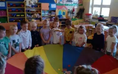 Dzieci stoją w sali przedszkolnej. Trzymają w rękach chustę animacyjną.