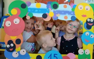 Dziewczynki trzymają w rękach ramkę ozdobioną kolorowymi kropkami. Zaglądają do niej. Uśmiechają się.