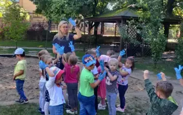 Dzieci stoją w ogrodzie przedszkolnym z nauczycielką. Na rękach mają niebieskie rękawiczki. Podnoszą ręce do g&oacute;ry. 