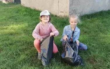 Dwie dziewczynki kucają na trawie. W rękach trzymają worki na śmieci. Na rękach mają rękawiczki. Uśmiechają się.