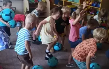 Dzieci są w sali przedszkolnej. Turlają piłkę stopami.