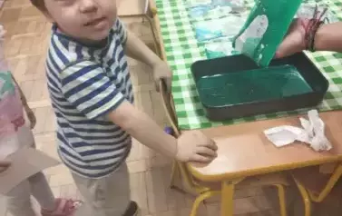 Chłopiec stoi przed stolikiem. Na stoliku stoi zielony pojemnik z farbą. Do pojemnika wkładana jest kartka.