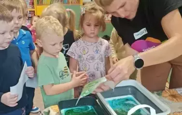 Dzieci stoją przy nauczycielce angielskiego. Przyglądają się. Pani wkłada do zielonego pojemnika kartkę. Macza ją w farbie.