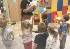 Dzieci stoją w sali przedszkolnej. Przed nimi stoi nauczycielka angielskiego. Pokazuje kartkę z obrazkiem.