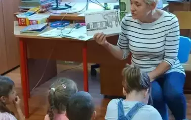 Dzieci siedzą na dywanie. Przed nimi siedzi Pani Bibliotekarka. Trzyma w ręku ilustrację. Tłumaczy dzieciom.
