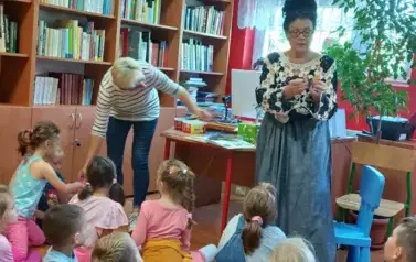Dzieci siedzą na podłodze. Przed nimi stoi bibliotekarka.