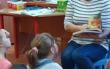 Dzieci siedzą przed Panią Bibliotekarką. Pani trzyma w ręku książkę.