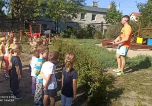 Dzieci stoją w szeregu przed nauczycielem w ogrodzie przedszkolnym.