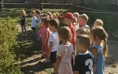 Dzieci stoją w szeregu w ogrodzie przedszkolnym. Patrzą przed siebie.