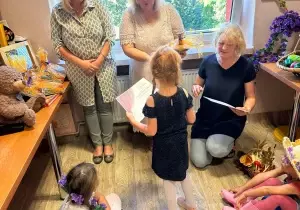 Pani Dyrektor i Wicedyrektor stoją, wręczają nagrodę i dyplom dziewczynce. Obok kuca nauczycielka. Trzyma w ręku kartkę. Na podłodze siedzą dzieci. Przyglądają się.