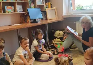 Dzieci siedzą na pufach w bibliotece przedszkolnej. Pani nauczycielka w okularach siedzi razem z dziećmi.