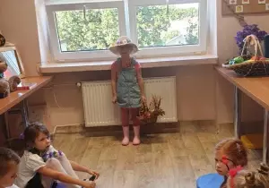 Dziewczynka stoi w bibliotece przedszkolnej. Przebrana jest za ogrodniczkę. Trzyma w ręku koszyk.
