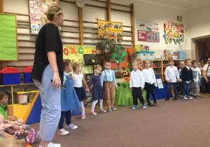 Dzieci stoją na środku sali. Obok nich stoi nauczycielka języka angielskiego.
