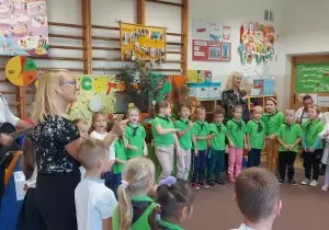 Dzieci z koła zuchowego "Leśne Ludki" stoją w zielonych koszulkach jeden obok drugiego. Razem z nimi stoją nauczycielki prowadzące.
