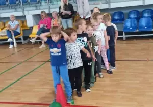 Dzieci stoją na sali gimnastycznej jeden za drugim. Przed nimi stoi czerwony stożek.