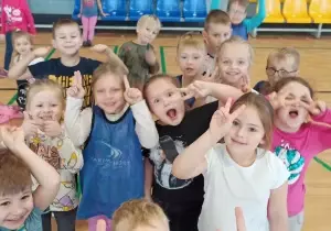 Dzieci machają do fotografa. Uśmiechają się. Robią śmieszne miny. Są na sali gimnastycznej.