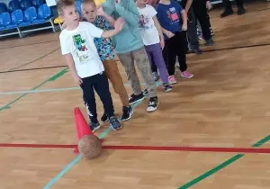 Dzieci stoją na sali gimnastycznej. Stoją jeden za drugim.