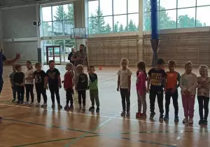 Dzieci stoją w szeregu. Przed nimi chodzi trener. Tłumaczy co będą robili podczas zajęć.