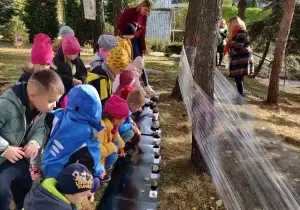 Dzieci stoją w lasku w ogrodzie przedszkolnym. Przed nimi wisi folia rozwieszona między drzewami.