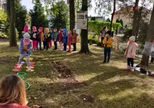 Dzieci wykonują zadanie sportowe. Stoją jeden za drugim. Pierwsza dziewczynka stoi na kolorowych talerzykach.