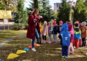 Dzieci stoją jeden obok drugiego. Stoją w lasku. Patrzą na nauczyciela. Nauczyciel trzyma w jednym ręku pacynkę łosia..