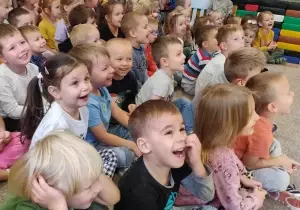 Dzieci siedzą na dywanie. Oglądają przedstawienie teatralne. Śmieją się.