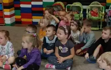 Dzieci siedzą na dywanie w sali przedszkolnej.