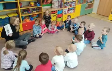 Dzieci siedzą na dywanie wkole. Wśr&oacute;d nich siedzi pani Zuzanna. Trzyma książkę. Opowiada.