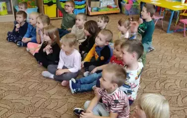 Dzieci siedzą na dywanie. Jeden chłopczyk podnosi rękę. Zgłasza się.