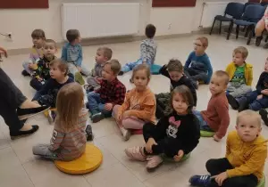 Dzieci siedzą na pufach w bibliotece. Uśmiechają się.