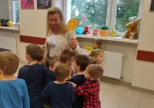 Pani nauczycielka stoi na środku. Trzyma w ręku ż&oacute;łtą, uśmiechniętą buźkę. Dzieci stoją przed nauczyceilką. Przytulają się do niej.