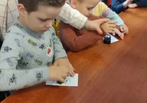 Dzieci stemplują datownikami na kartkach. Pani trzyma jednego chłopca za ręke. Pomaga mu stemplować.