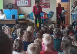 Dzieci siedzą tyłem na dywanie. Przed nimi stoi pan. M&oacute;wi do nich przez mikrofon.
