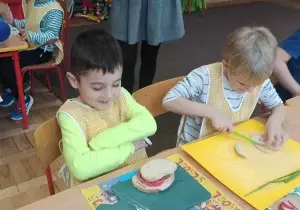Dw&oacute;ch chłopc&oacute;w siedzi przy stole. Jeden z nich uśmiecha się i patrzy na przygotowaną kanapkę. Drugi kroi nożem na desce. kanapkę.