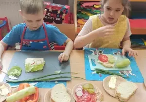 Dziewczynka z chłopcem siedzą przy stoliku. Na sobie mają fartuszki. Dziewczynka trzyma w ręku kanapkę. Chłopiec ozdabia swoją kanapkę.