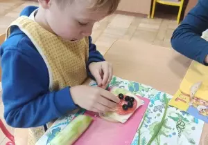 Chłopiec ozdabia kanapkę. Ubrany jest w fartuszek. Siedzi przy stoliku.