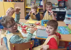 Dzieci siedzą przy stoliku. Ubrane są w fartuszki. Uśmiechają się do zdjęcia. Na stoliku leżą wykonane kanapki.