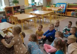 Dzieci siedzą na dywanie w siadzie skrzyżnym. Słuchają opowiadania czytanego przez mamę Zosi.