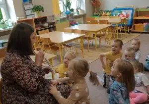 Dzieci siedzą na dywanie. Na krzesełku siedzi mama Zosi. Pokazuej skiążkę dzieciom. Dziewczynka opiera się o kolana mamy Zosi.