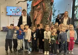 Grupa dzieci uśmiecha się. Za nimi stoją dwie nauczycielki. Za nimi stoi drzewo. Dzieci są w teatrze.