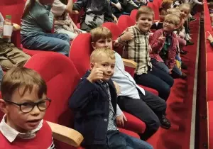 Dziecisiedzą w fotelach teatralnych. Uśmiechają się do zdjęcia. Ubrane są w koszule i sukienki.