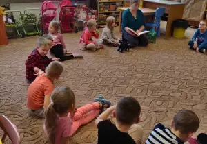 Dzieci wraz z rodziem siedzą na dywanie w sali przedszkolnej.