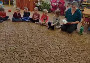 Dzieci wraz z rodziem siedzą na dywanie w sali przedszkolnej.
