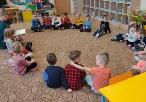 Dzieci sidzą w kole na dywanie. Razem z nimi siedzi rodzic.