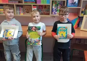 Trzech chłopc&oacute;w stoi w bibliotece przedszkolnej. Trzymają w ręku książki wygrane w ramach konkursu. Uśmiechają sie do zdjęcia.