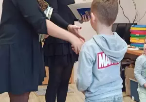 Chłopec otrzymuje książkę w ramach nagrody za konkurs. Nauczycielka wyciąga do niego rękę.