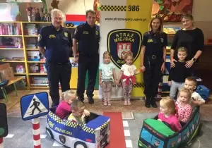 Zdjęcie grupowe dzieci ze Strażą Miejską. Z tyłu stoi baner Straży Miejskiej. Dzieci stoją na makiecie przejścia dla pieszych.